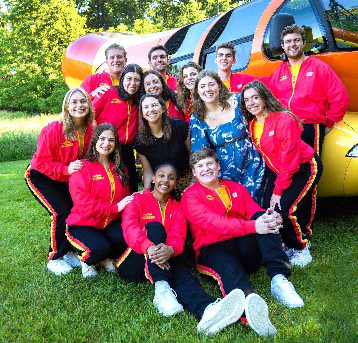 Out of more than 7,000 applicants for the 2024-25 season, Oscar Mayer selected these 12 Wienermobile drivers, posing with Wienermobile coordinator Emily Schmitt (blue dress) and head dog Molle Twing (black dress) in Madison, Wisconsin, where the drivers trained for two weeks in June 2024.