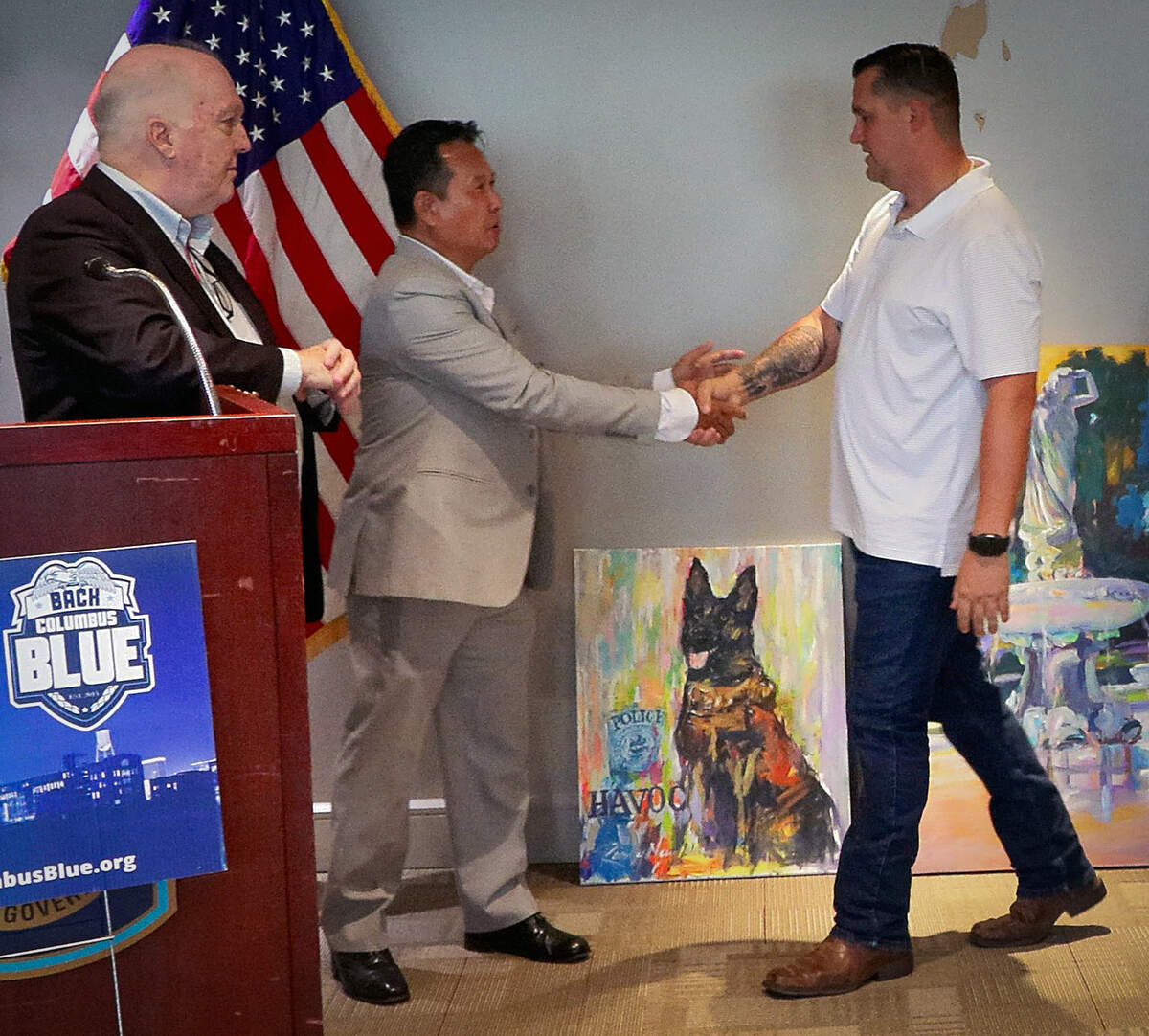 K9 officer Havoc’s handler, Cpl. Brent Wilson of the Columbus Police Department, right, greets Columbus artist Randy Nguyen, center, at a June 29, 2026, news conference as Jed Harris, chairman and co-founder of Back Columbus Blue, looks on.