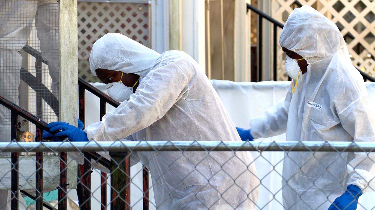 Workers scrape recently paint from a railing at a house. The work area is fenced off and bears warning signs telling residents it’s a “lead work area.”