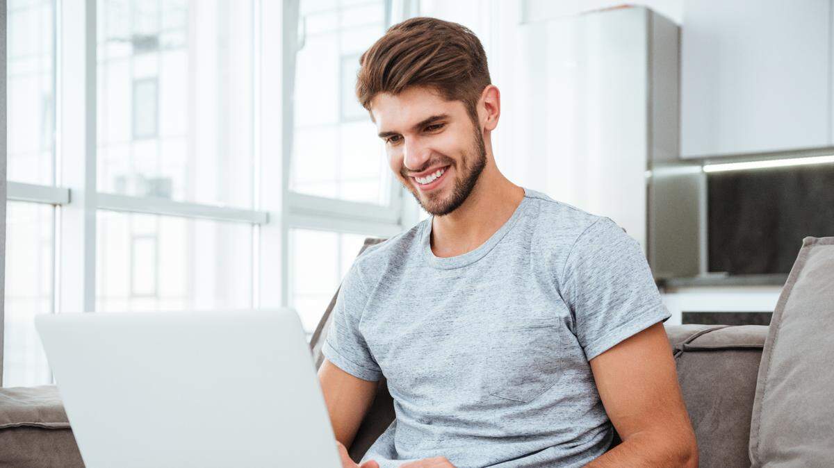 Happy young man in t-shirt sitting on sofa at home. Working on laptop computer and smiling.